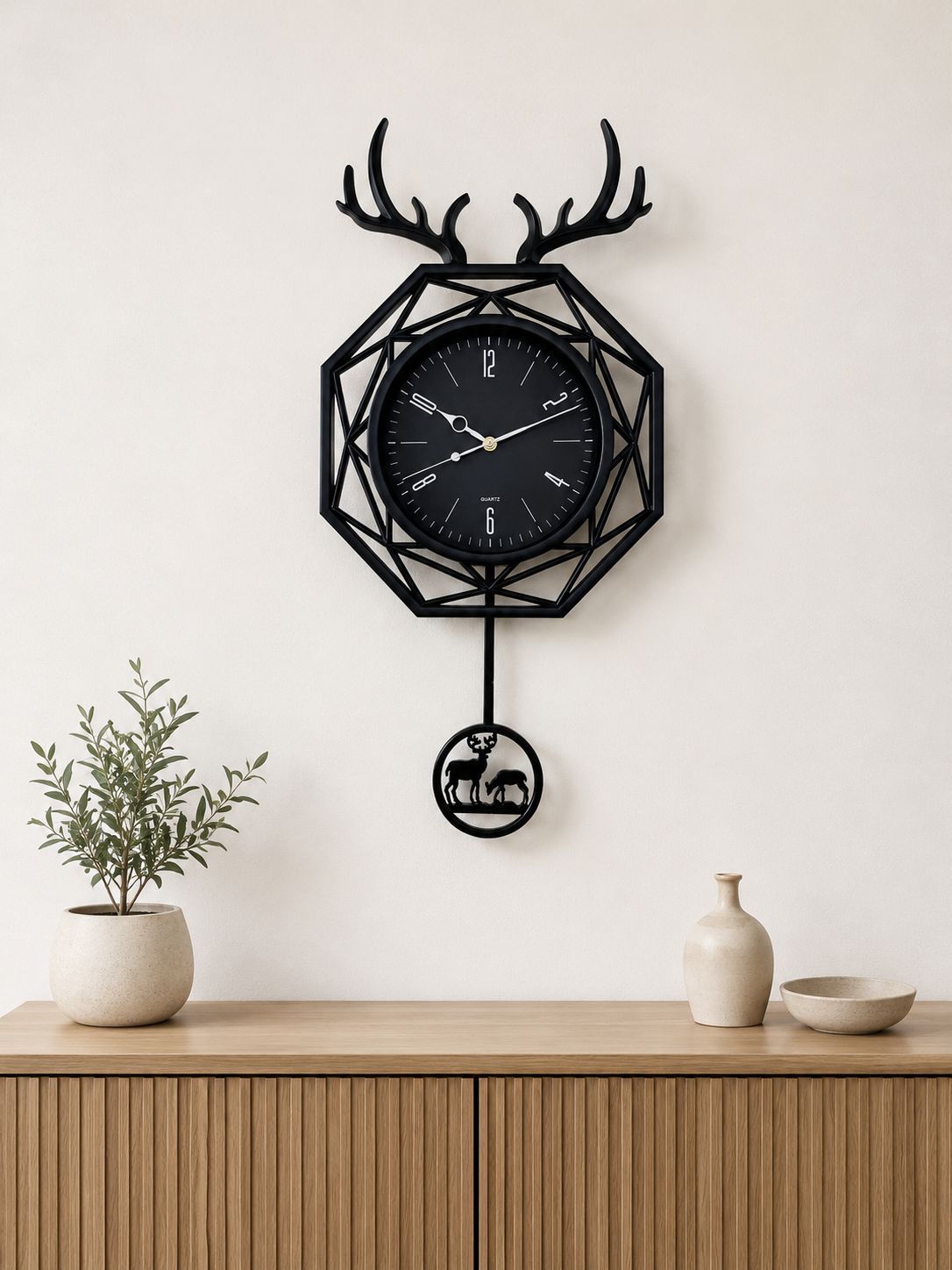 A black metal geometric wall clock with antler motifs at the top and a hanging deer figure below, displayed on a light wall above a wooden console table with a potted plant and ceramic vases.