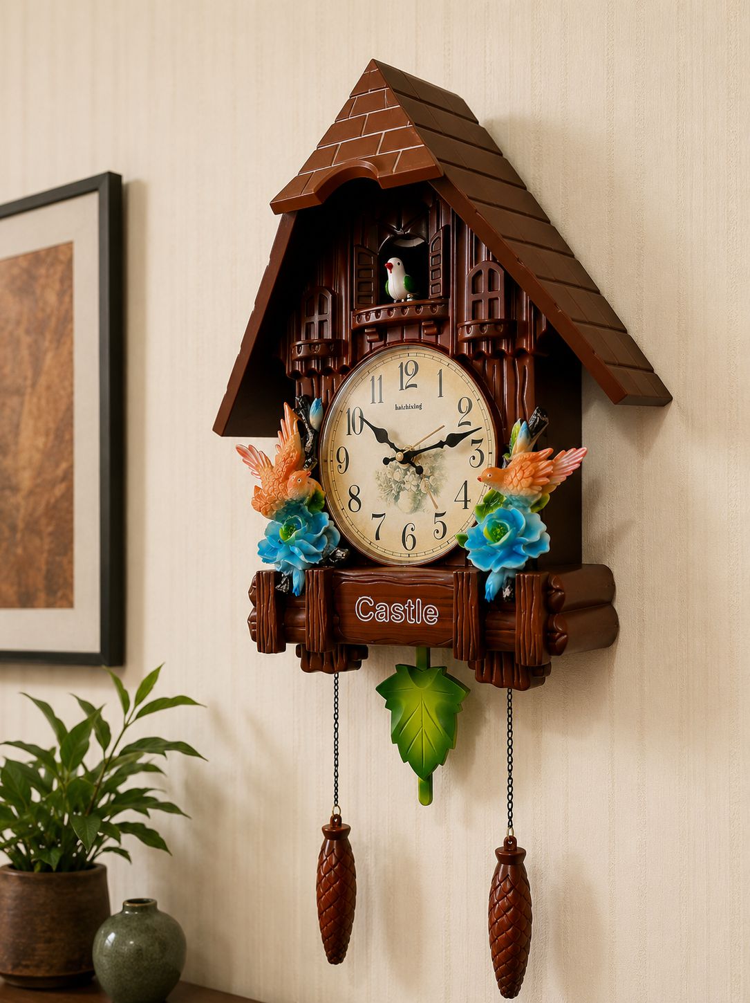 A brown cuckoo clock shaped like a wooden house with a shingled roof, cream clock face, decorative flowers, and hanging pine-cone weights, mounted on a beige wall in a home interior.
