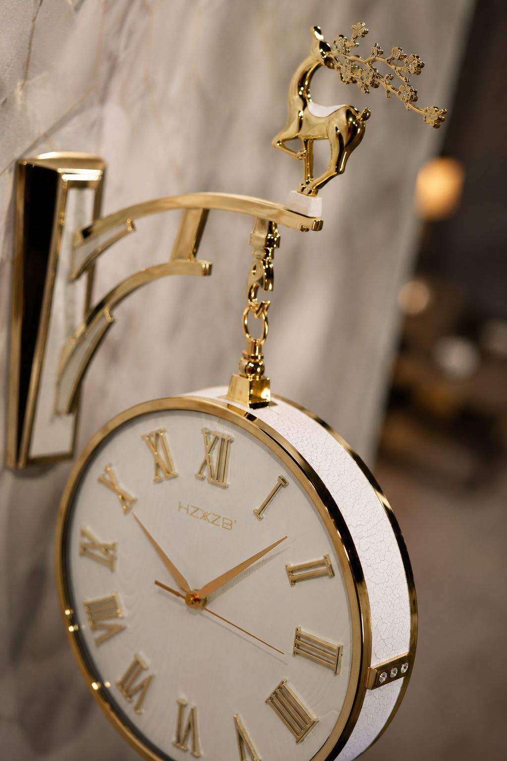 A close-up of a decorative wall clock with a white dial, gold Roman numerals, and a gold metal bracket and deer ornament, mounted against a softly blurred marble wall.
