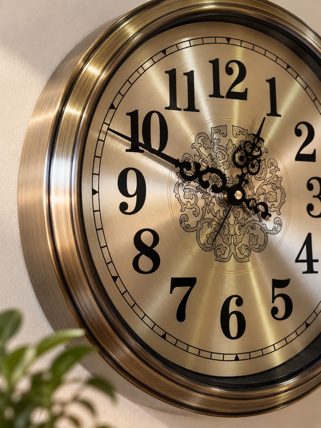 A close-up of a round wall clock with a brushed brass or bronze-toned frame and a metallic beige clock face featuring large black Arabic numerals and ornate black hands.