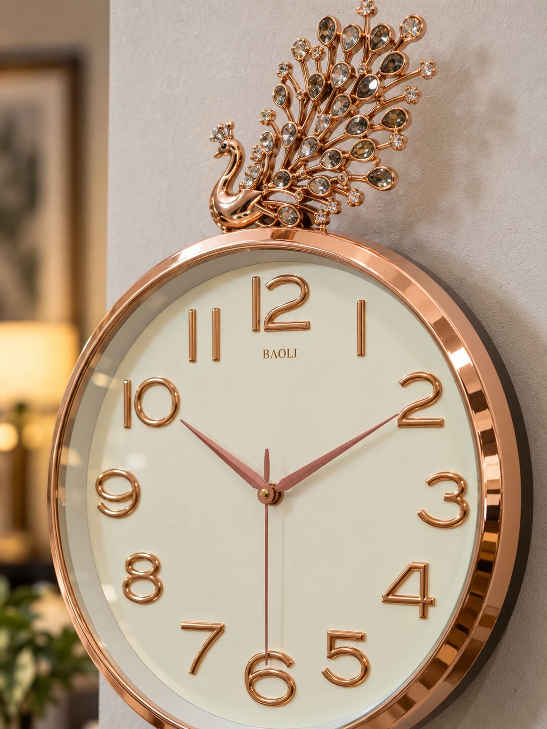 A close-up of a wall clock with a rose gold peacock topper and cream-colored dial, mounted on a light gray wall.