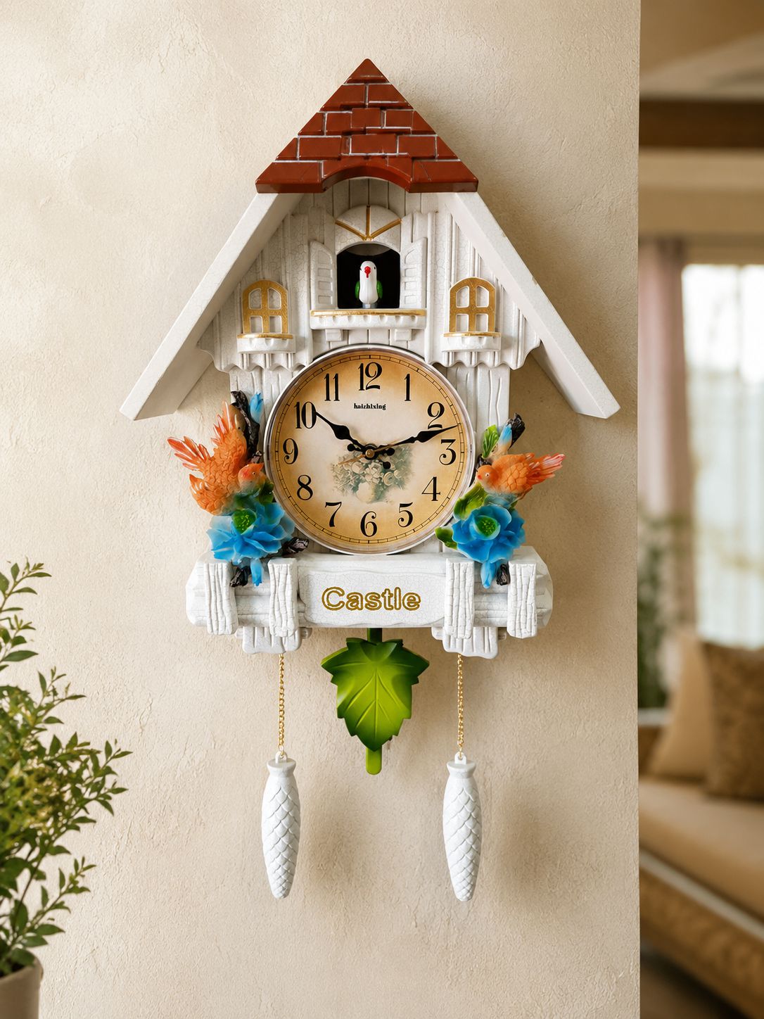 A cuckoo-style wall clock shaped like a small white house with a brown brick roof, gold-toned clock face, decorative flowers, and two hanging white weights, mounted on a beige wall in an indoor room.