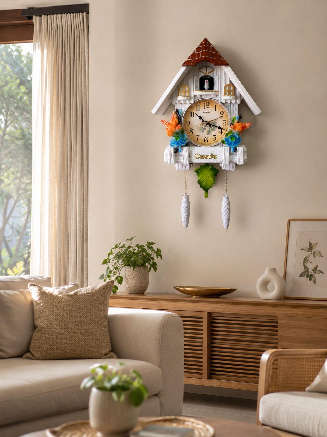 A cuckoo clock shaped like a white cottage hangs on a beige wall in a styled living room. The clock has a red roof, a cream-colored face, and colorful bird details in orange, blue, and green, with white dangling weights below.