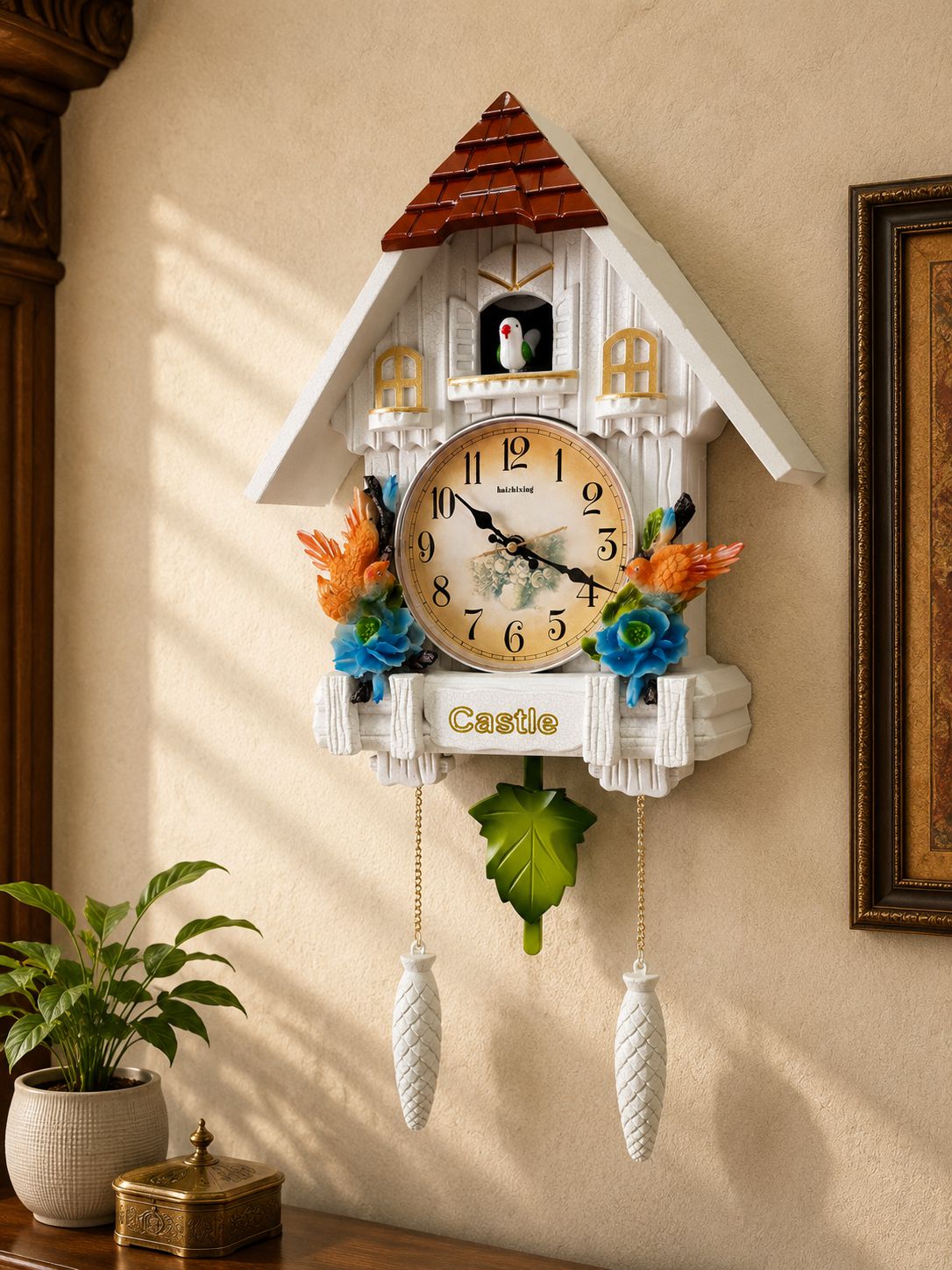 A cuckoo clock shaped like a white cottage with a brown roof, mounted on a beige wall. The clock face is cream with black numerals, decorated with orange and blue flowers, a green leaf, and two white hanging weights.