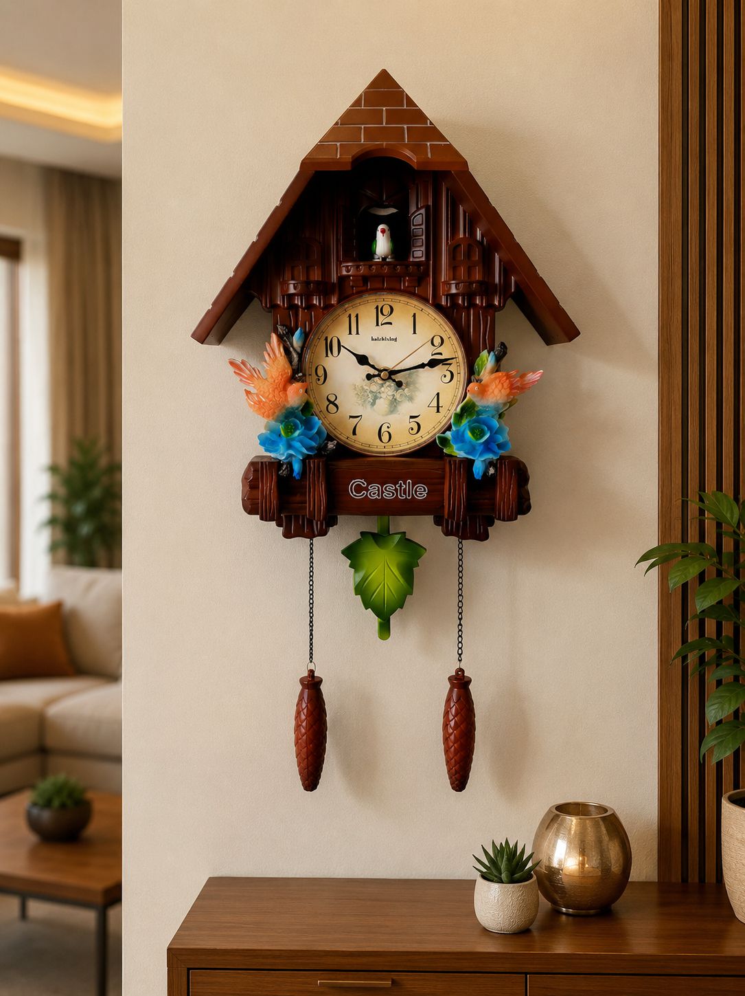 A cuckoo clock styled like a traditional wooden chalet house, mounted on a beige wall in a living room setting. The clock features a brown wood-grain body, a cream-colored clock face with black numbers, colorful artificial flowers in orange, blue, and green, and two dangling brown pendulums.