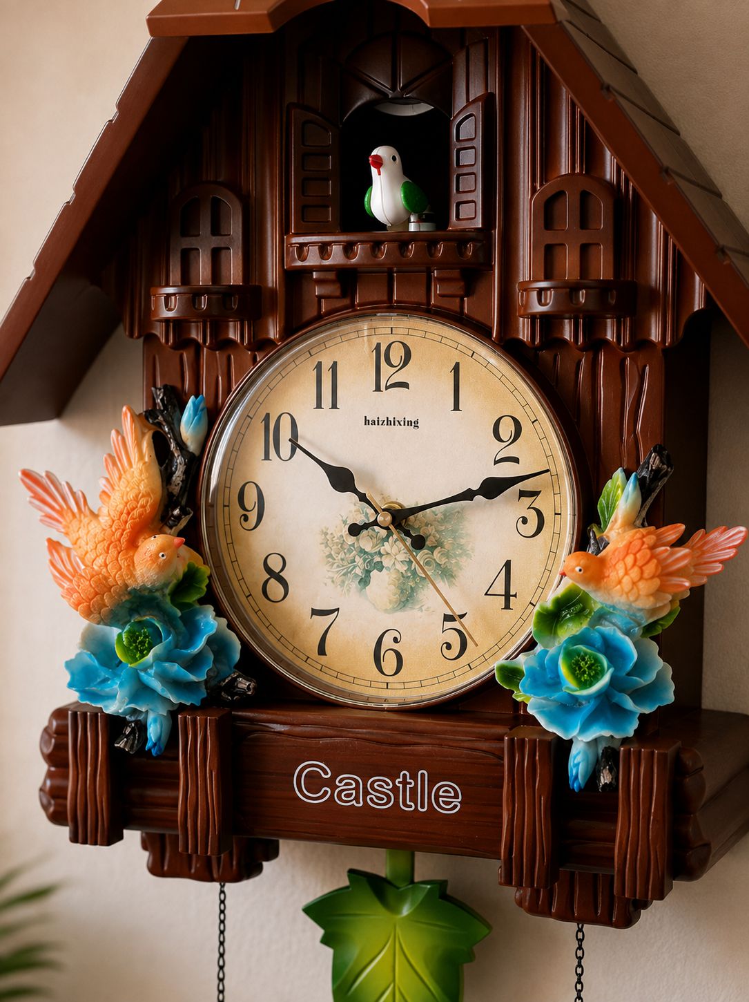 A decorative cuckoo-style wall clock shaped like a brown wooden house, with a round cream clock face, black numerals, ornate bird and flower ornaments in orange, blue, and green, and the word "Castle" on the lower front.