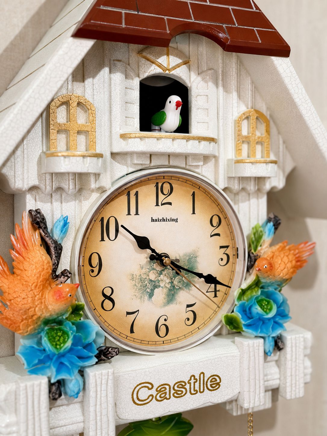A decorative cuckoo-style wall clock shaped like a white castle with a brown roof, floral accents, and two small birds in windows. The clock face is cream with black numerals and hands, and the word 'Castle' appears at the bottom.