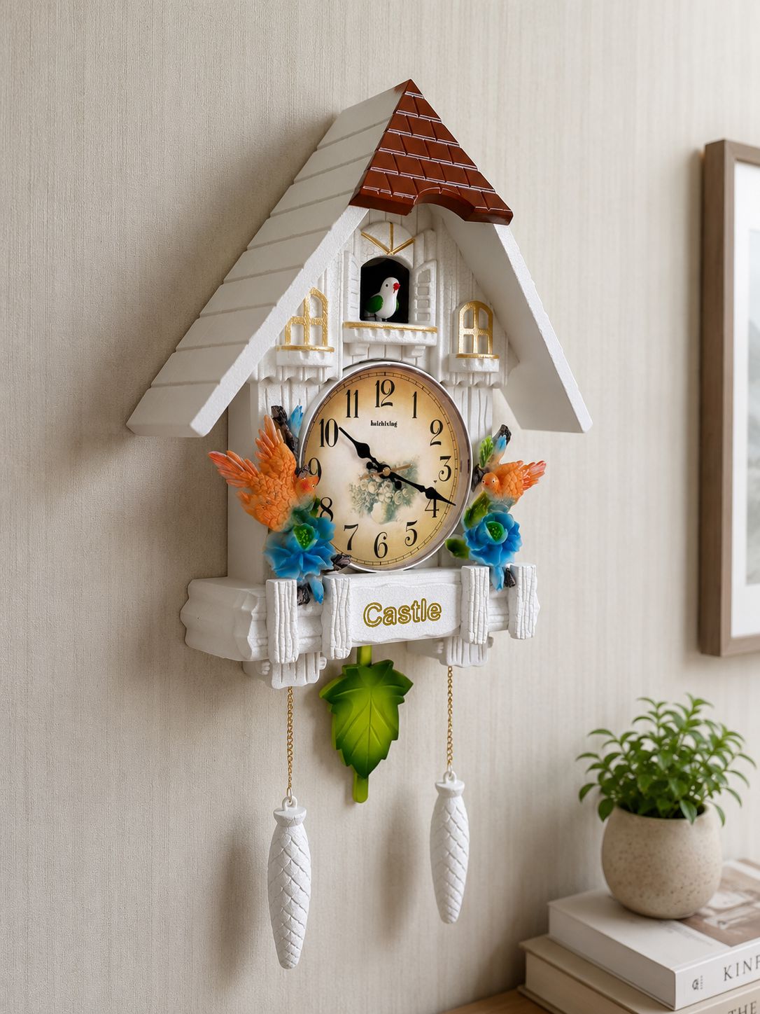 A decorative cuckoo clock shaped like a white cottage with a brown roof, mounted on a beige wall. The clock features colorful bird and flower accents, hanging white weights, and a round clock face with black numerals.