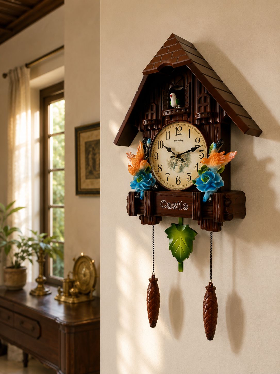 A decorative cuckoo clock shaped like a wooden house hangs on a beige wall in a sunlit room. The clock has a cream-colored face with black numerals and hands, colorful floral accents on either side, a small bird in the window, and two hanging pine-cone weights below a green leaf.