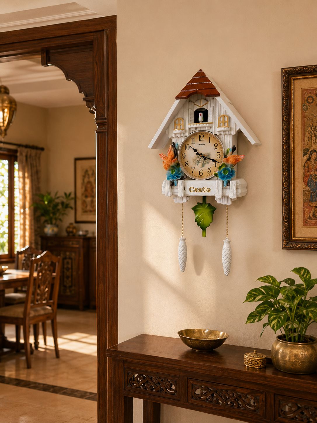 A decorative cuckoo clock styled like a white cottage with a red roof, mounted on a beige wall in a warm, traditional interior. The clock features colorful floral accents, a green cuckoo bird, and hanging white weights.