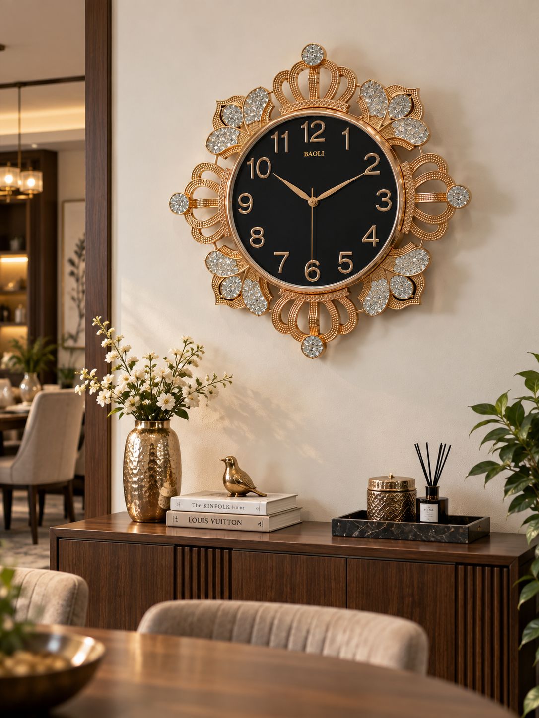 A decorative wall clock with a black face and ornate gold frame, mounted on a beige wall above a wooden console table in a styled interior setting.