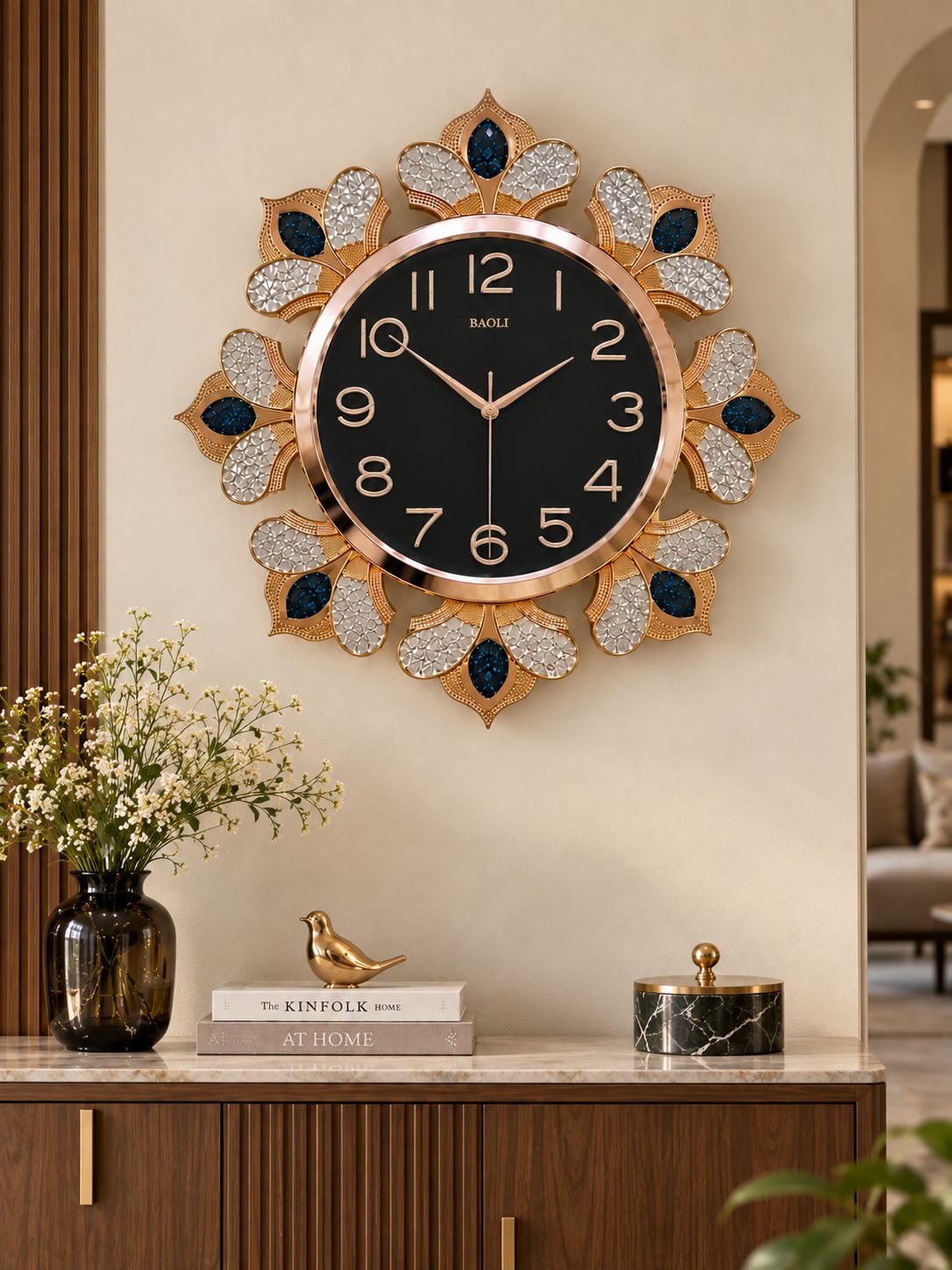 A decorative wall clock with a black face, gold-tone numerals and hands, and a gold-toned sunburst frame embellished with rhinestones and dark blue teardrop accents. The clock is mounted on a beige wall above a wooden console table with a vase of flowers and decorative objects.
