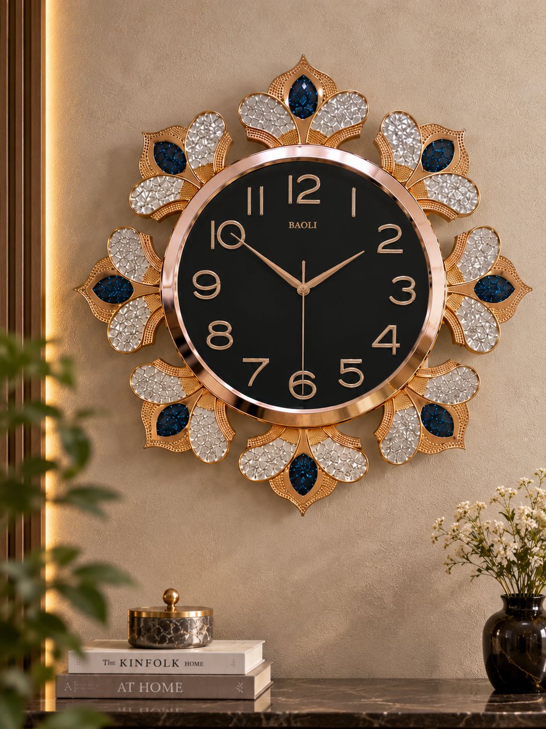 A decorative wall clock with a black face, gold-tone numerals and hands, and a sunburst frame made of gold, clear rhinestones, and dark blue teardrop accents. The clock is mounted on a beige wall above a console table with books and flowers.