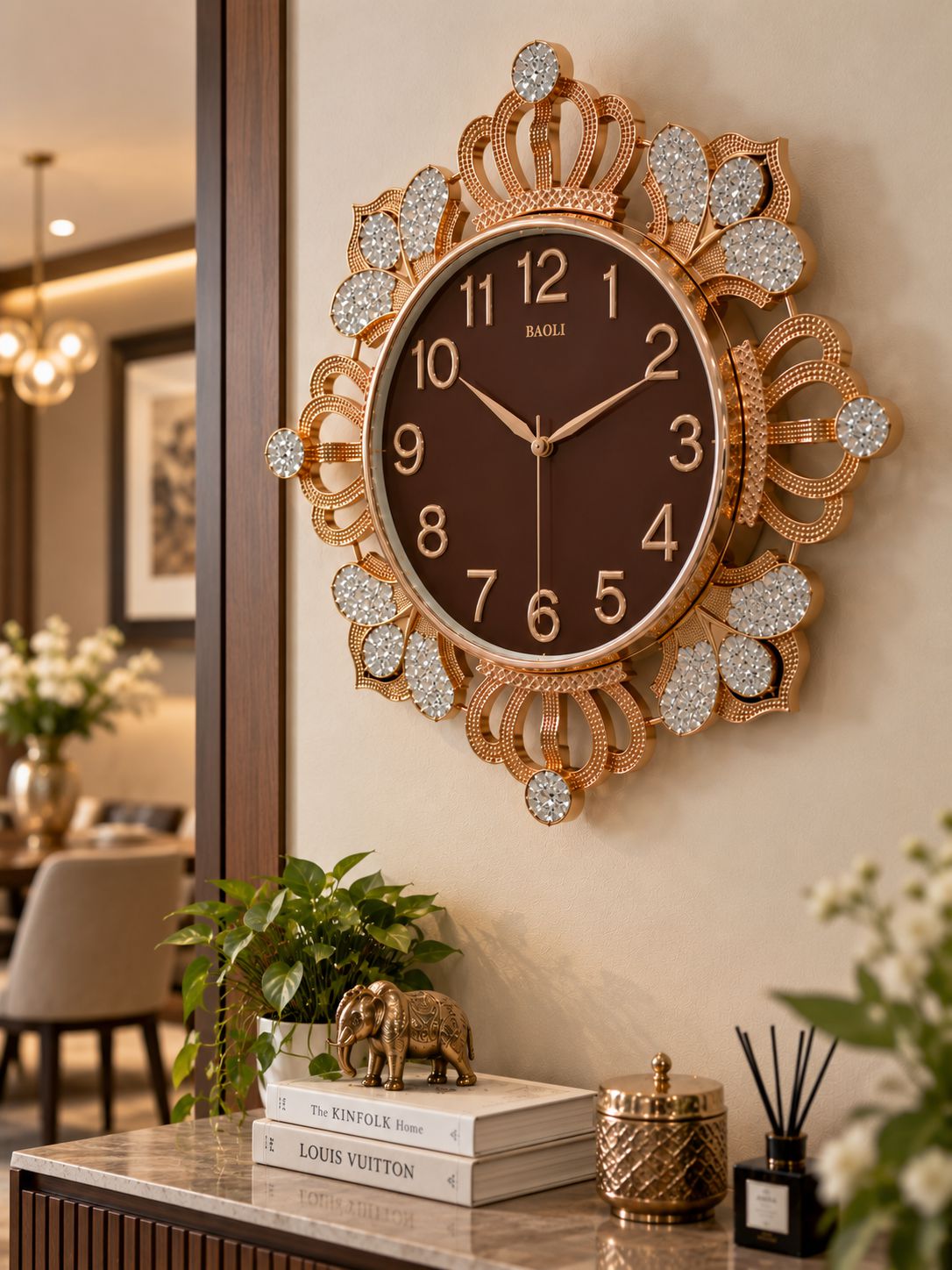 A decorative wall clock with a brown face and ornate gold-tone frame embellished with rhinestone-like accents, mounted on a beige wall above a styled console table in a warm interior setting.