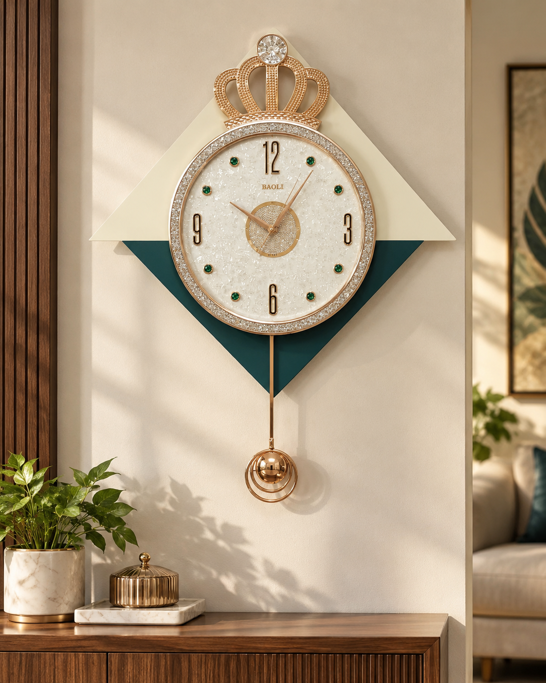 A decorative wall clock with a crown motif and a hanging pendulum, mounted on a cream wall above a wooden console. The clock face is white with black numerals and gold hands, surrounded by a crystal-like border with small green accents.