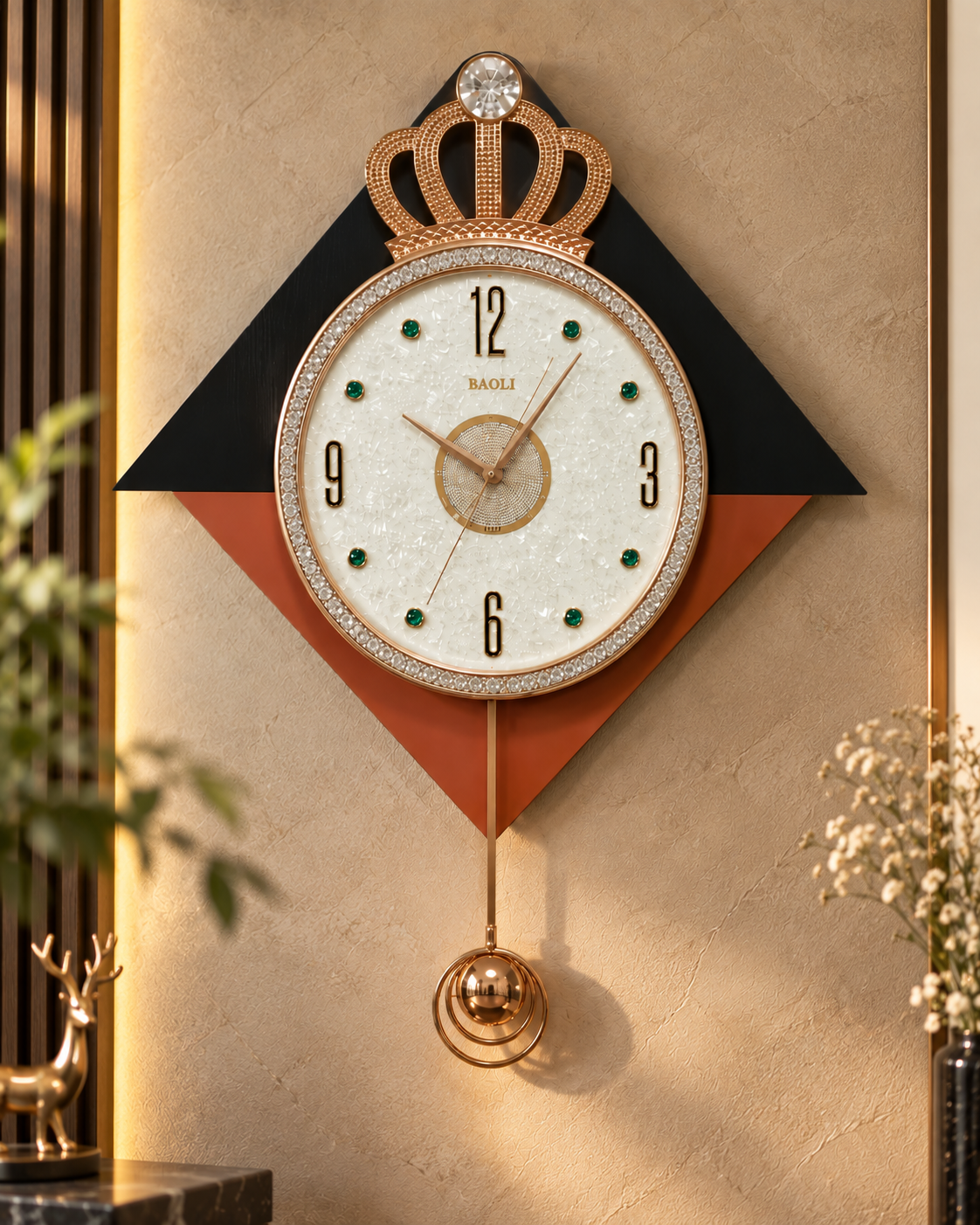 A decorative wall clock with a crown motif and hanging pendulum, mounted on a beige wall. The clock features a white textured dial with black numerals, a gold-toned center, green dot accents, and a rhinestone-embellished bezel.