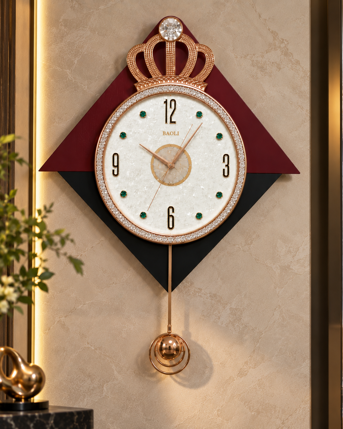 A decorative wall clock with a crown motif at the top, a white oval face, black Arabic numerals, green dot hour markers, and a hanging ball pendulum. The clock is mounted on a beige marble-look wall with warm lighting, flanked by dark geometric panels.