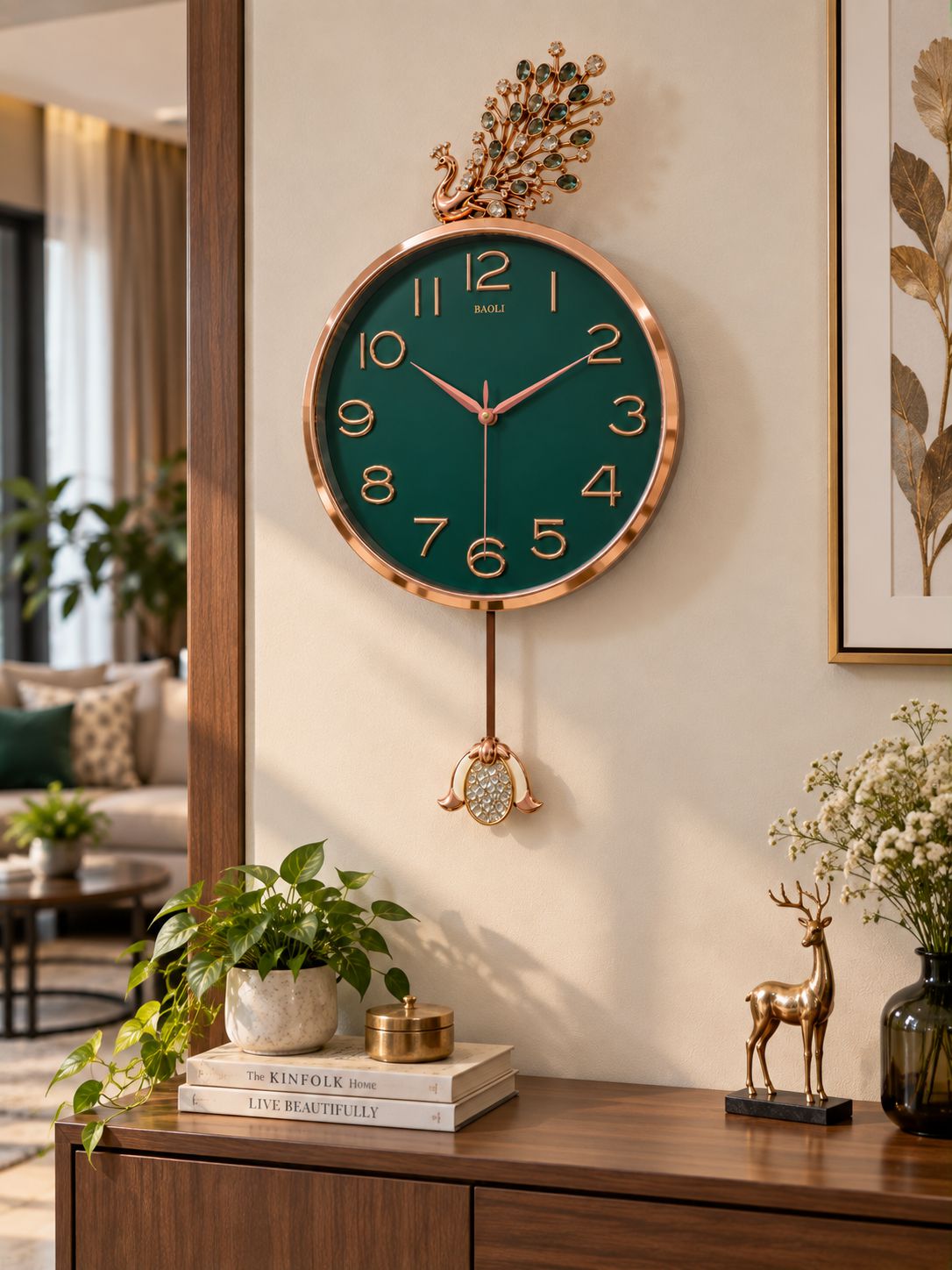 A decorative wall clock with a dark green face, rose gold-toned frame and hanging ornament, mounted on a beige wall above a wooden console. The room includes plants, books, a deer figurine, and framed wall art.