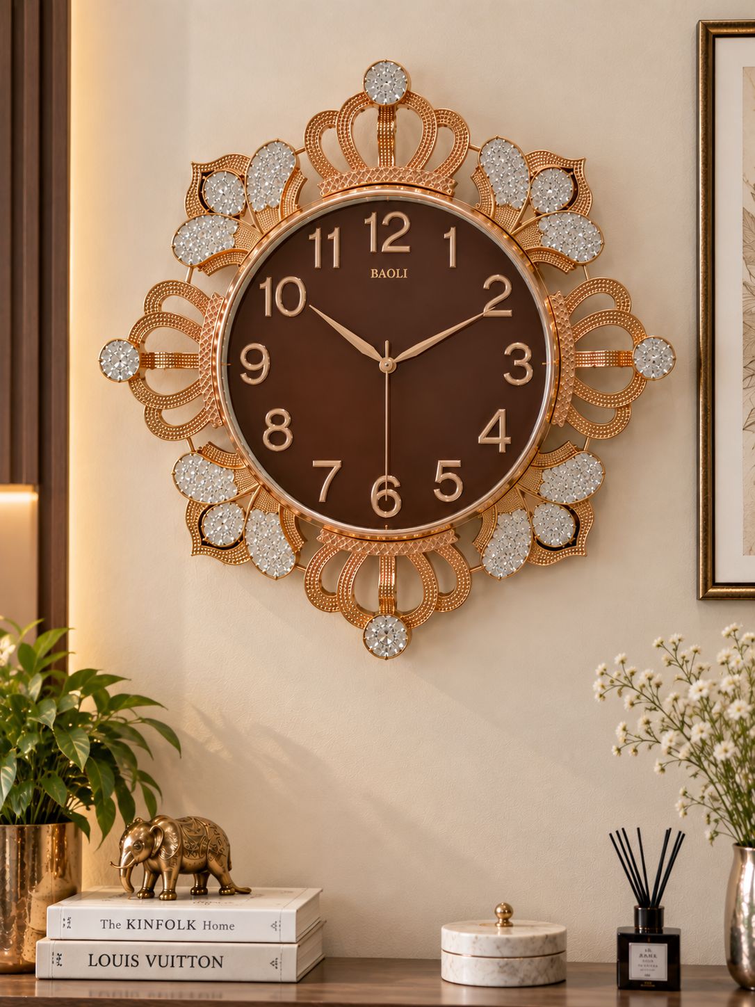 A decorative wall clock with a gold-toned ornate frame, crystal-like embellishments, and a dark brown clock face, mounted on a beige wall above a styled tabletop with books, a plant, and flowers.