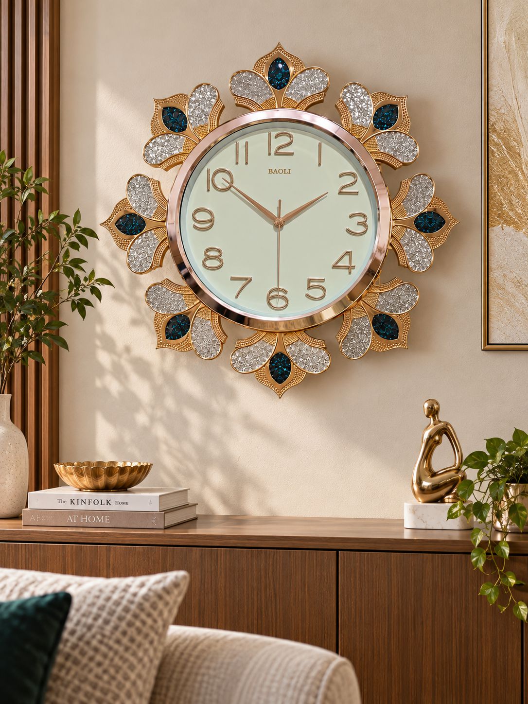 A decorative wall clock with a gold-toned sunburst frame, crystal-embellished petal shapes, and dark blue jewel accents, mounted on a beige wall above a wooden console in a styled interior.