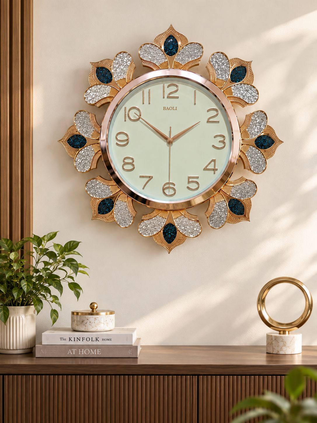 A decorative wall clock with a gold-toned sunburst frame, silver glitter petal details, and dark blue jewel accents, mounted on a beige wall above a wooden console table with books and plants.