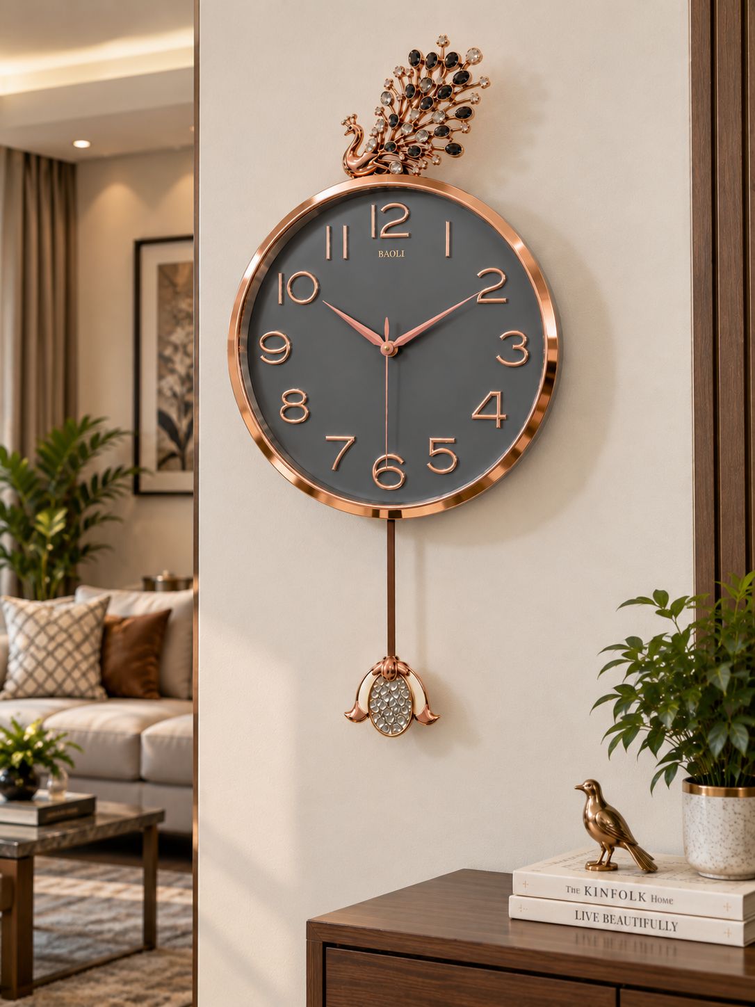 A decorative wall clock with a round dark gray face, rose gold-tone frame and numerals, a hanging pendulum with a crystal-encrusted ornament, and a peacock topper, shown mounted on a beige wall in a styled interior.