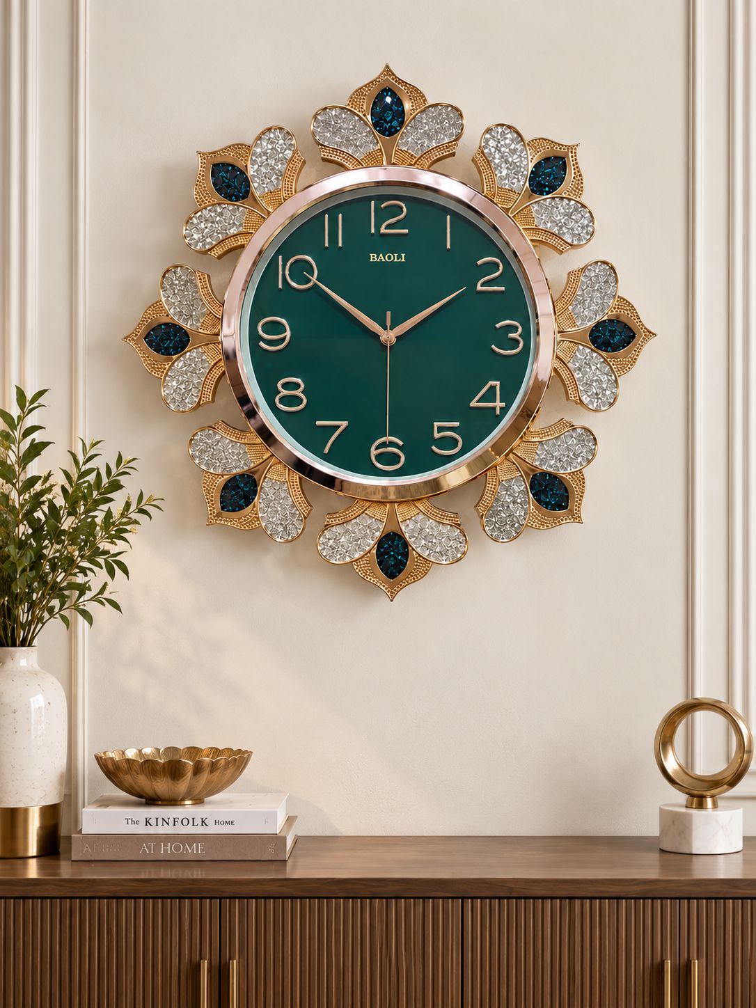 A decorative wall clock with a round green face and ornate gold-tone frame, surrounded by butterfly-like motifs with dark blue and silver rhinestone details. The clock is mounted on a cream wall above a wooden console table with books and decor.