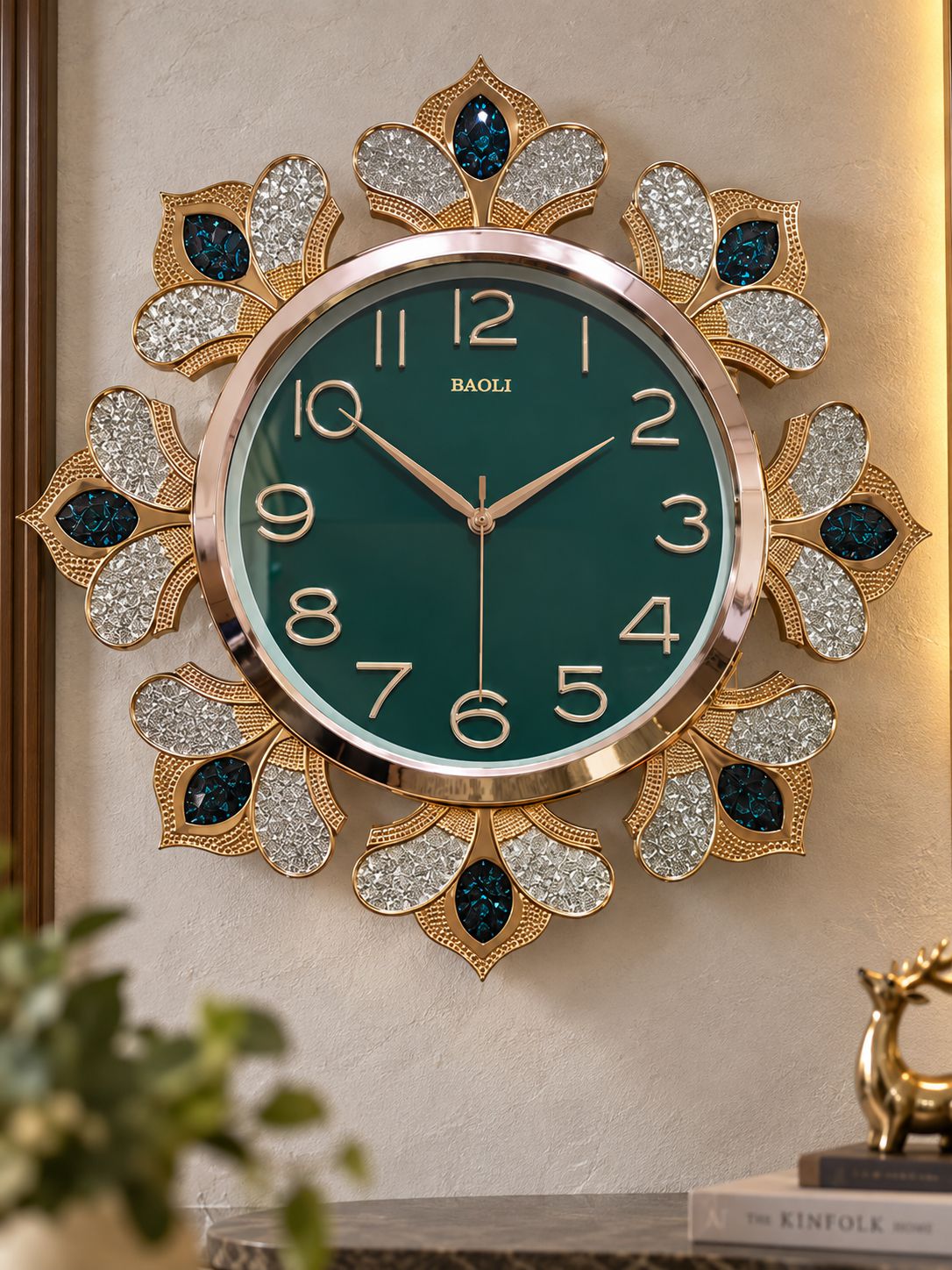 A decorative wall clock with a round green face, gold-tone numerals and hands, and an ornate floral frame adorned with rhinestones and dark blue teardrop accents, mounted on a beige wall.