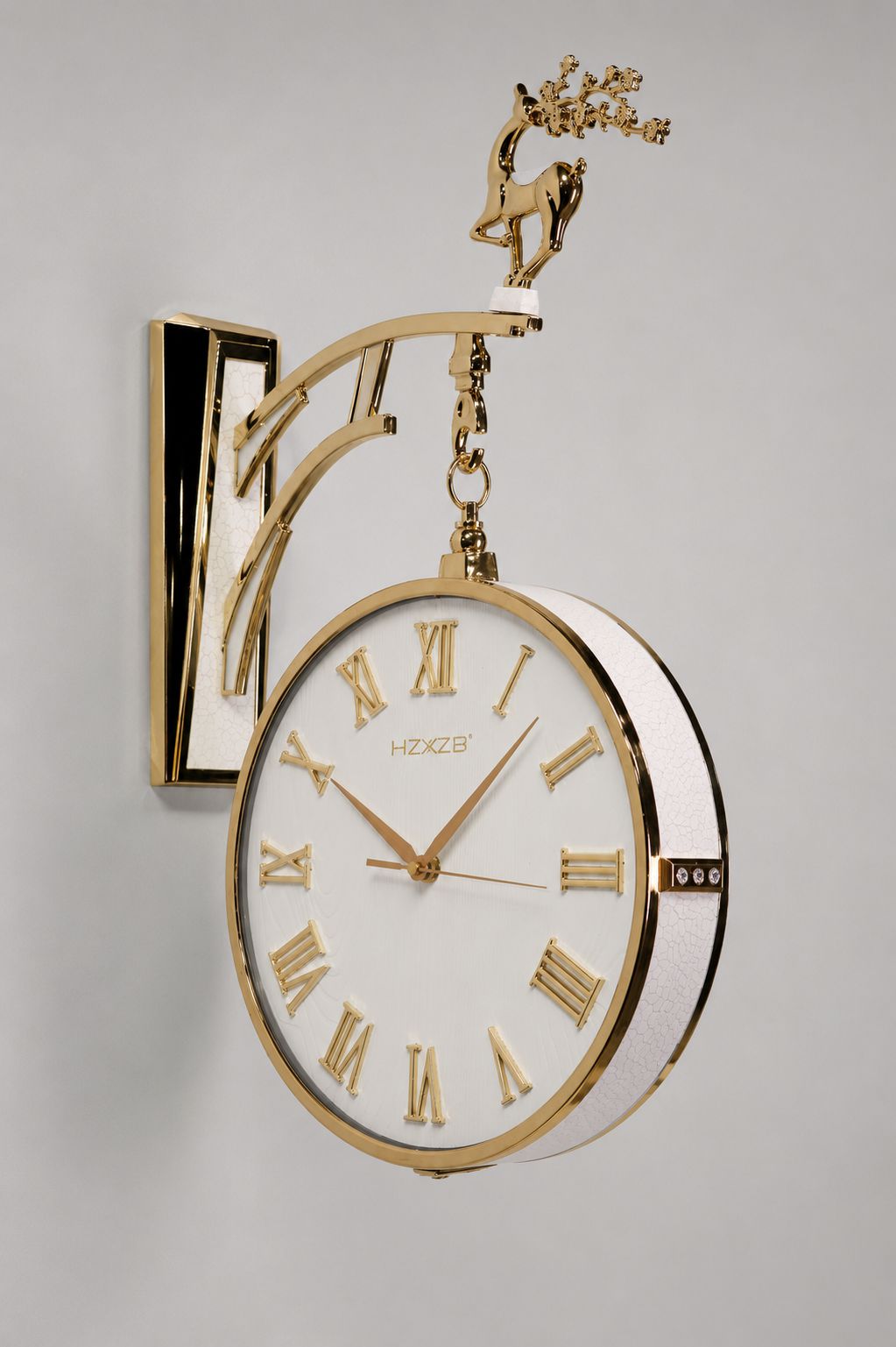A decorative wall clock with a white face, gold Roman numerals, and a polished gold metal bracket and frame. The bracket features a sculpted gold deer holding a flowering branch at the top.