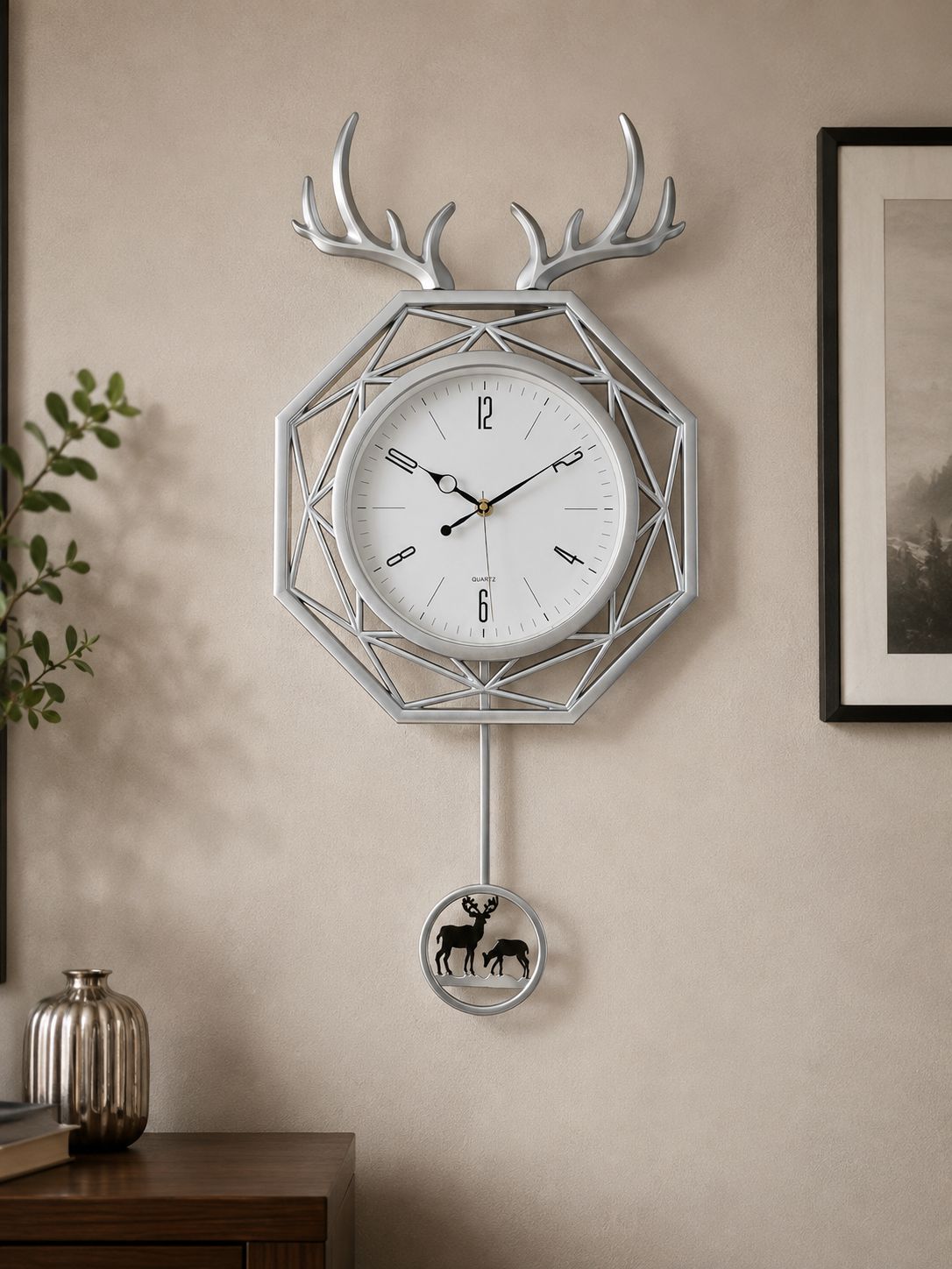 A geometric metal wall clock with deer antlers and a hanging circular ornament featuring a deer silhouette, mounted on a beige wall in a styled interior setting.