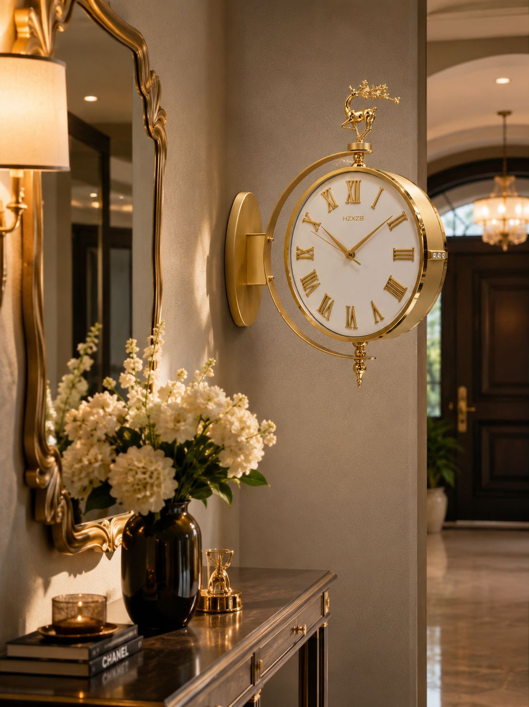 A gold-framed wall clock with a white face and Roman numerals mounted on a beige wall in an elegant interior hallway. A dark console table below holds white flowers in a black vase, a mirror, and decorative objects.