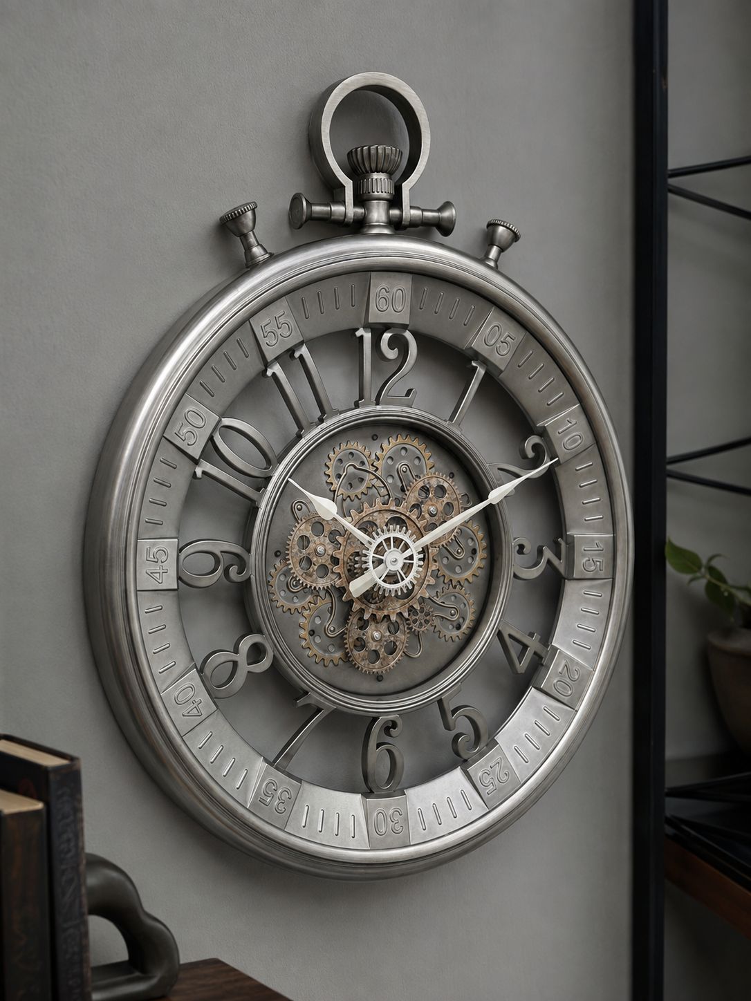 A large round wall clock styled like a pocket watch, with a metallic silver outer frame, exposed inner gears, and ornate black Arabic numerals, mounted on a gray wall.