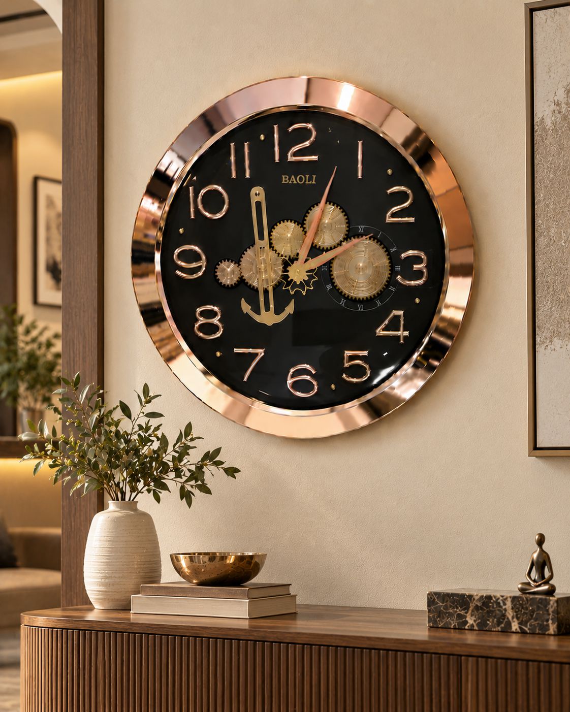 A large round wall clock with a black face, gold numerals and hands, and a rose-gold metallic frame, mounted on a beige wall above a wooden console table with decorative items.
