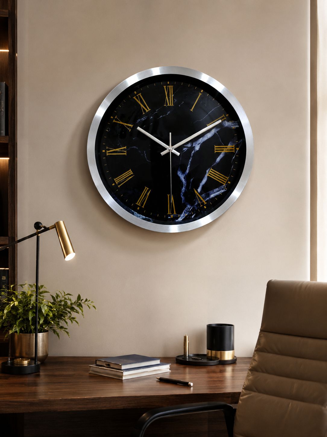 A large round wall clock with a black marble-patterned face and gold Roman numerals, mounted on a beige wall above a wooden desk with a lamp, books, and a chair.