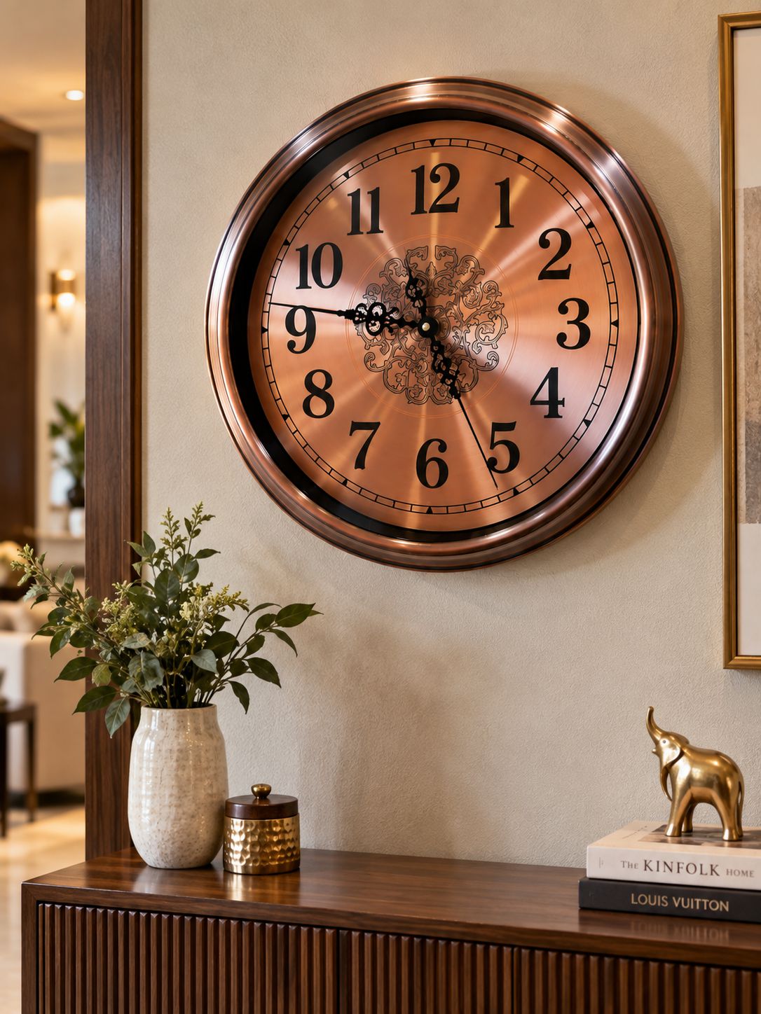 A large round wall clock with a bronze-toned metal frame and a copper-gold gradient dial, featuring black Arabic numerals and ornate hands. The clock is mounted on a beige wall above a dark wood console table decorated with a white vase of greenery, a small gold container, and a gold elephant figurine.