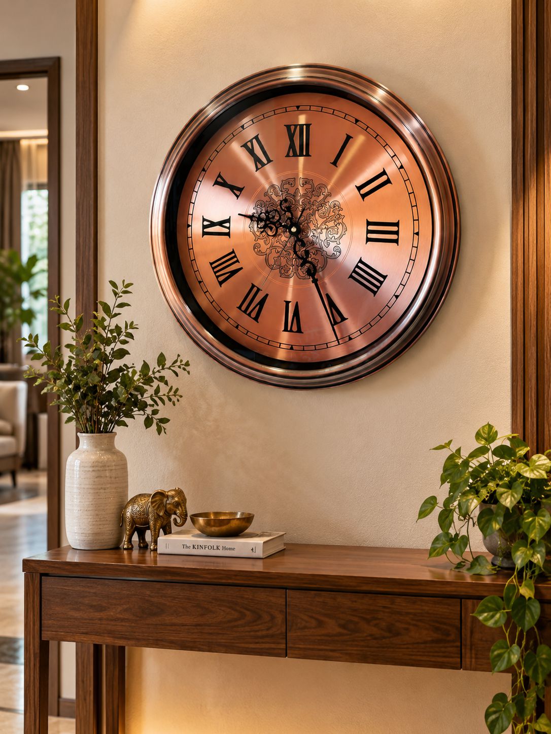 A large round wall clock with a bronze metallic finish and Roman numerals, mounted on a beige wall above a wooden console table decorated with plants and small figurines.