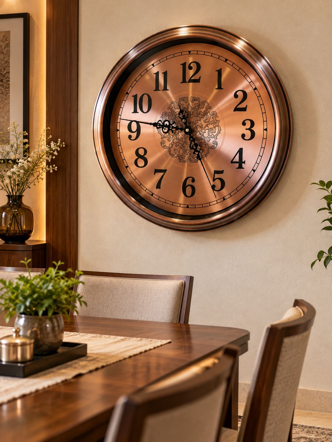 A large round wall clock with a bronze metallic frame and a warm copper-toned face, mounted on a beige wall above a wooden dining table with chairs.