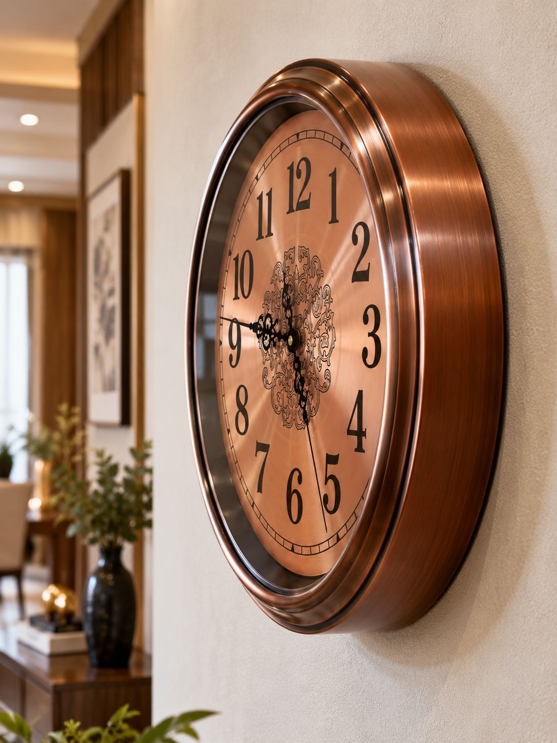 A large round wall clock with a brushed copper-brown frame and a matching clock face featuring black Arabic numerals and ornate black hands, mounted on a light beige wall in a home interior setting.