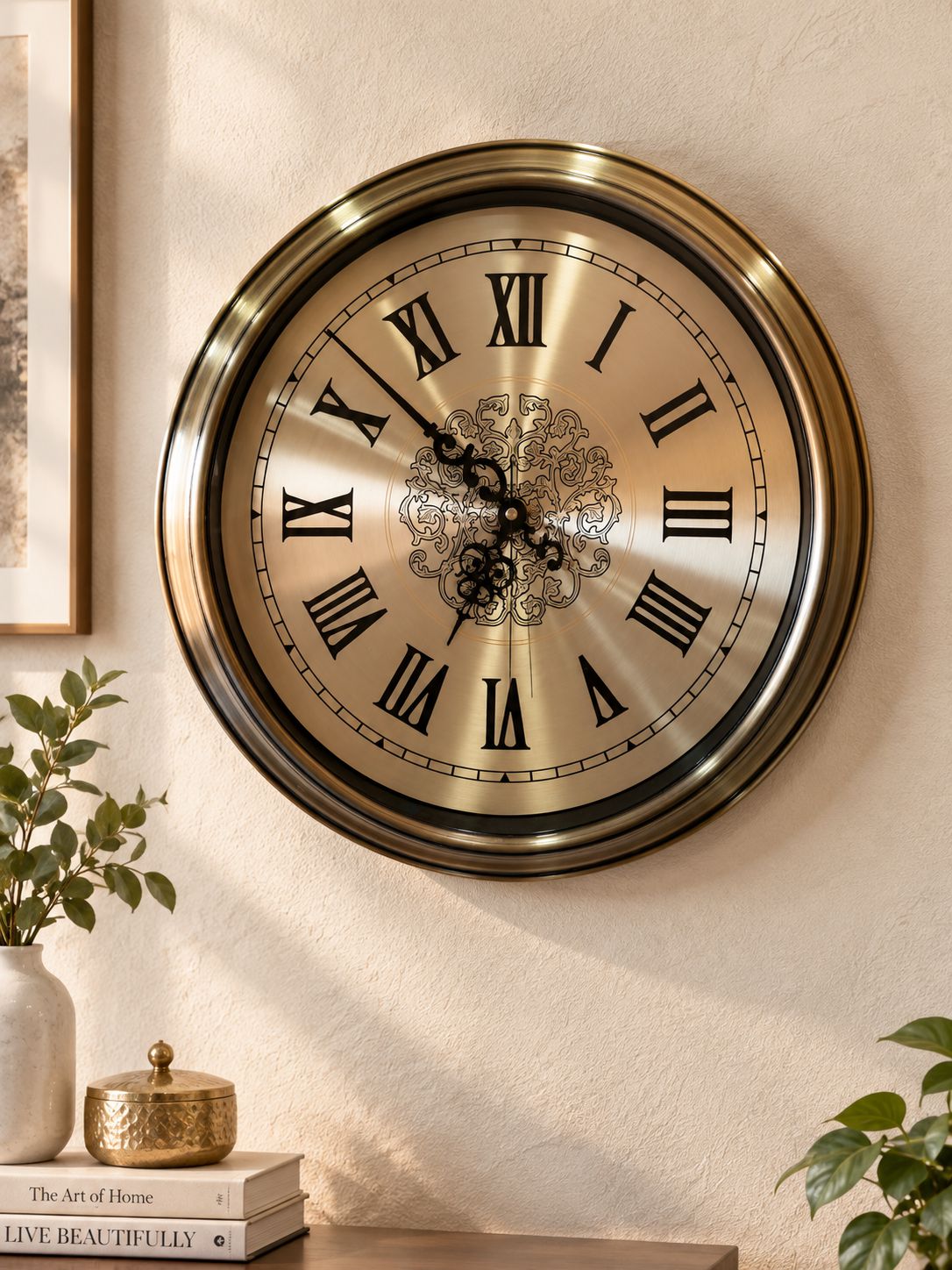 A large round wall clock with a brushed gold-toned metal frame and Roman numerals, mounted on a beige wall above a tabletop with books and plants.