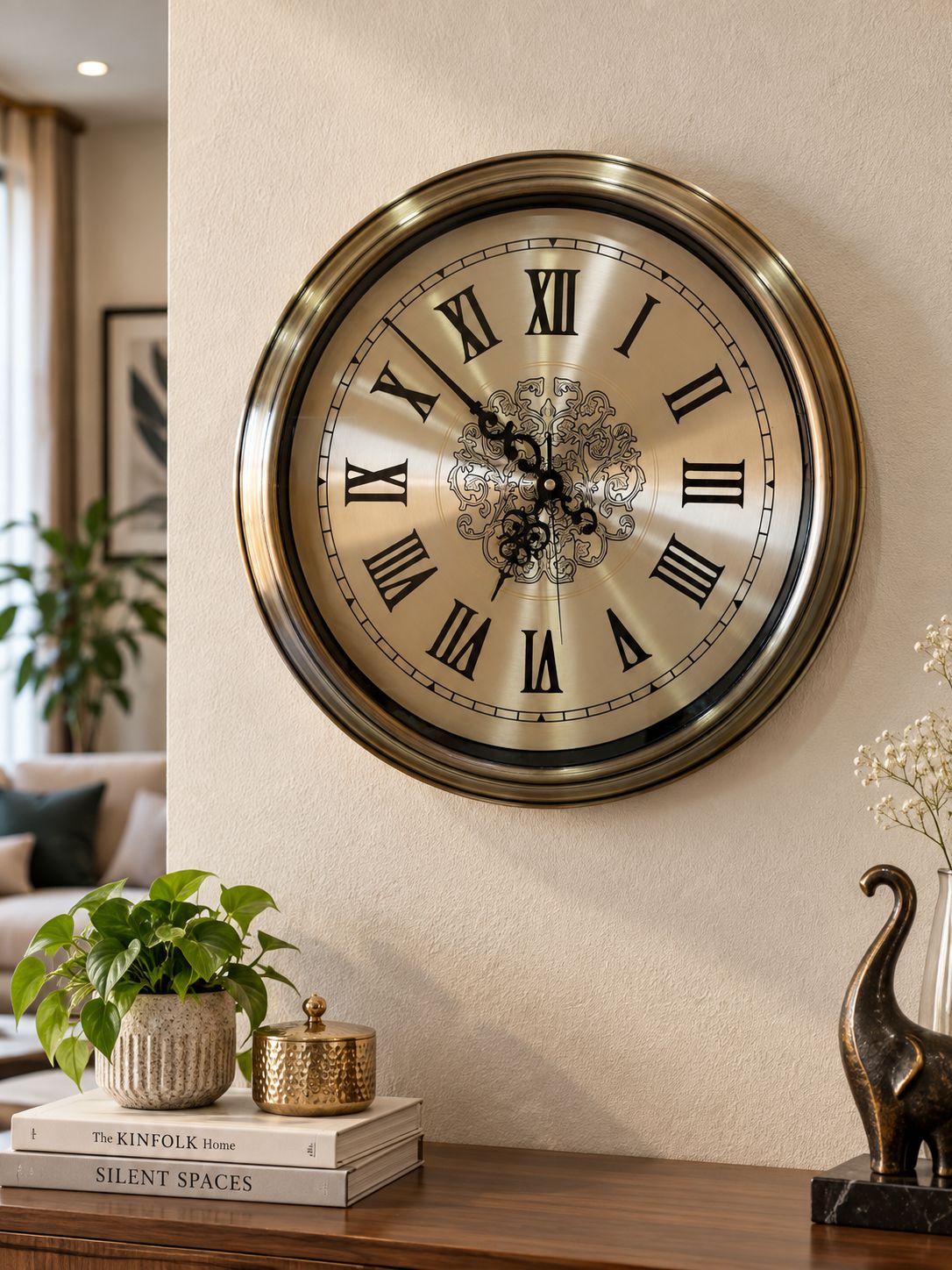 A large round wall clock with a brushed gold-toned metal frame and Roman numerals, mounted on a beige wall above a wooden console table with books, a plant, and decorative objects.