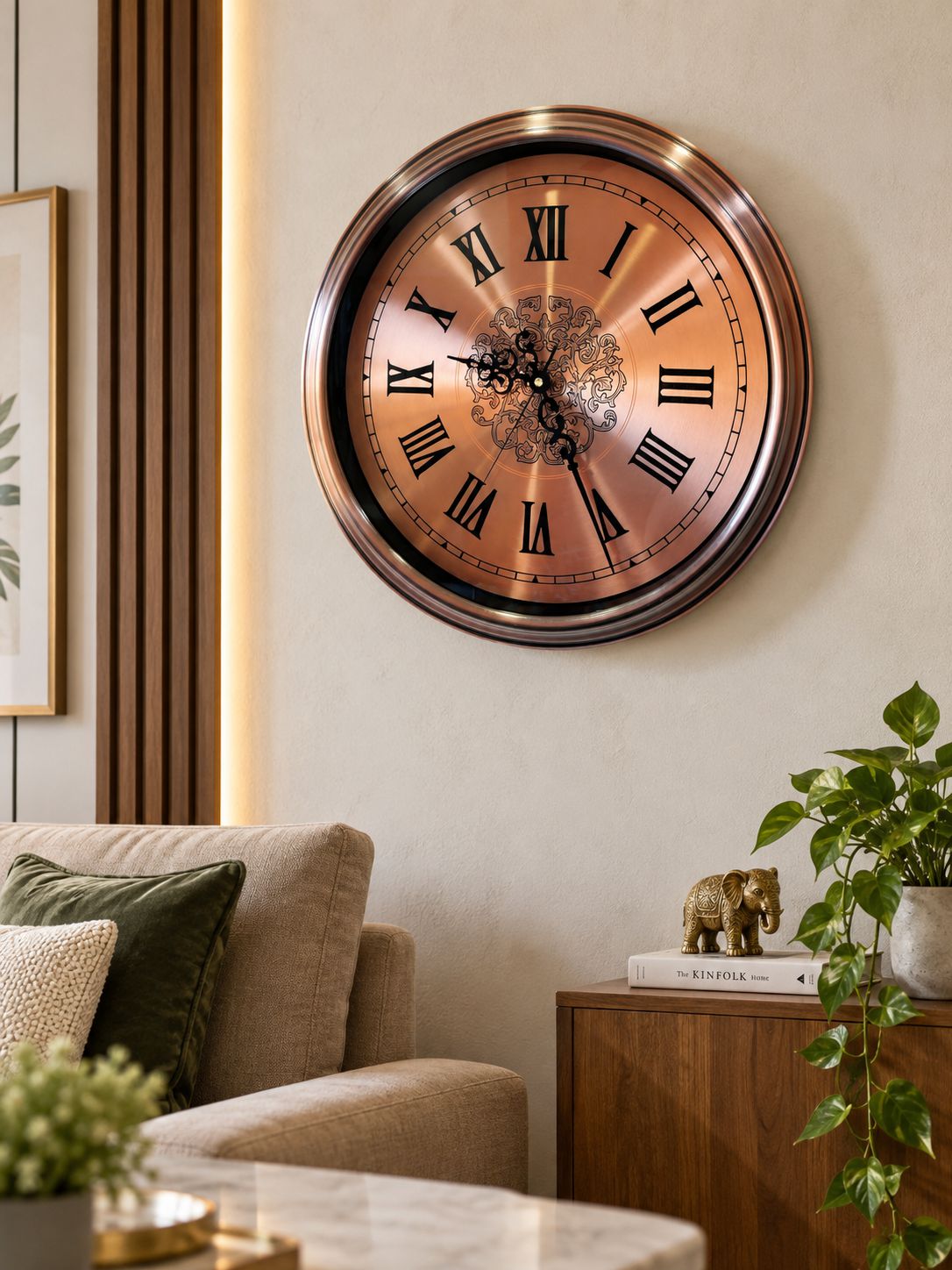 A large round wall clock with a copper-toned face and black Roman numerals, mounted on a beige wall in a modern living room setting with a sofa, side table, and green plant.