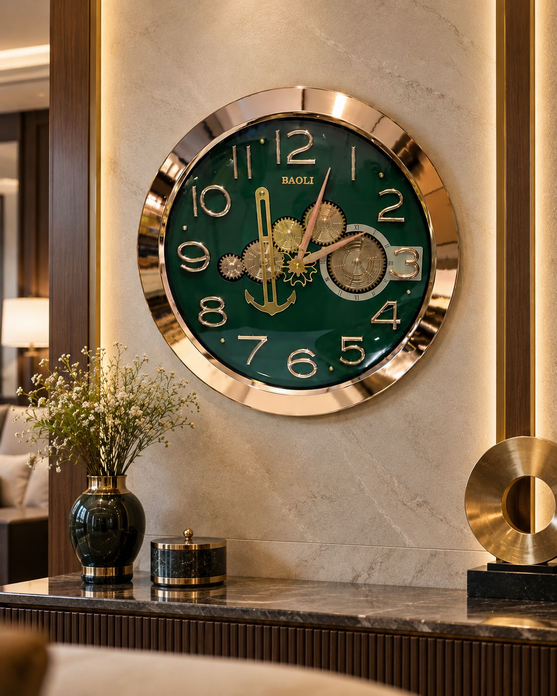 A large round wall clock with a dark green face, gold-tone numerals and hands, and exposed mechanical gears in the center, mounted on a beige marble wall above a console table with decorative vases and a gold sculpture.