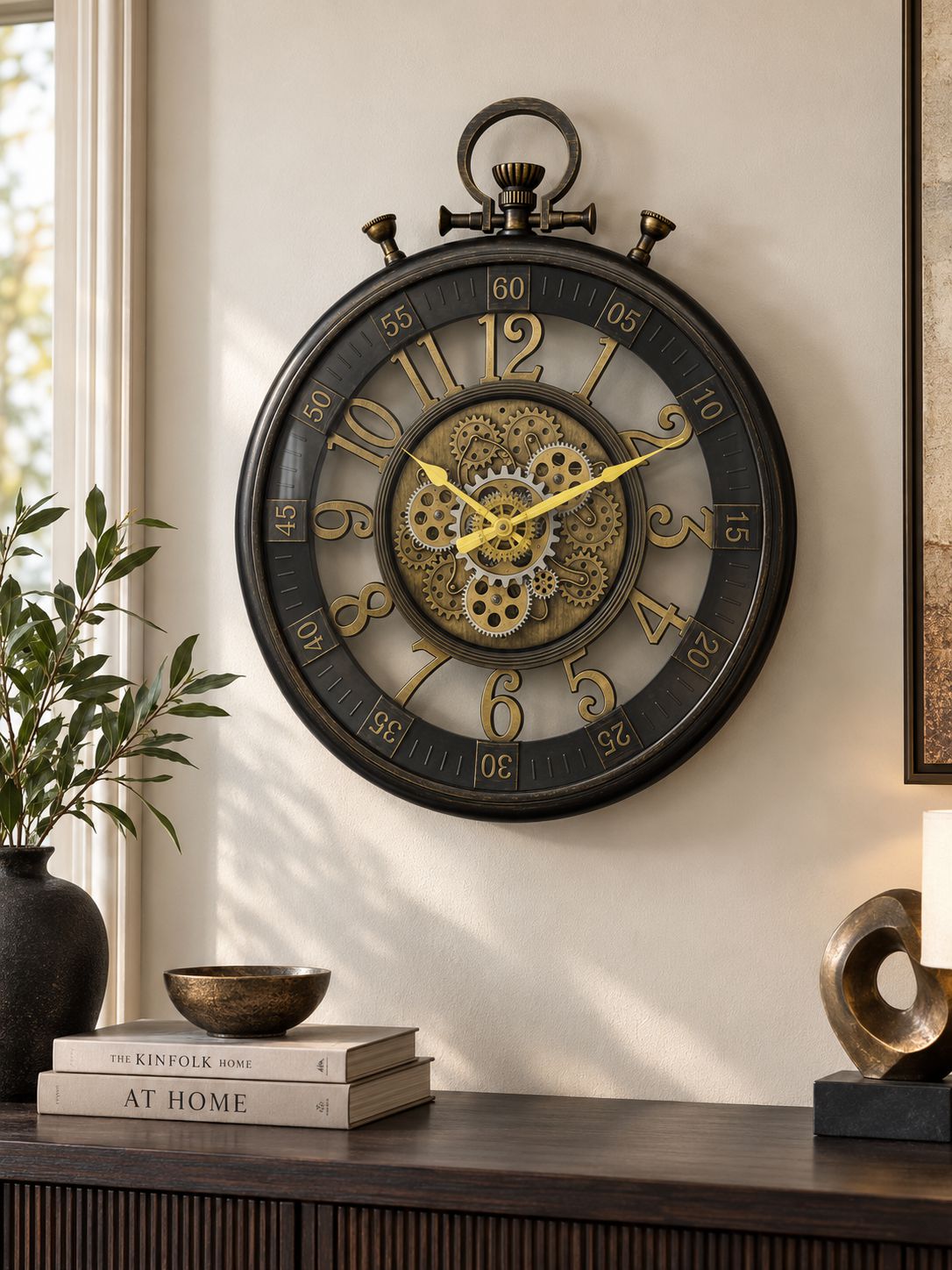 A large round wall clock with a dark metal frame and exposed inner gears, mounted on a beige wall above a wooden console table with books and decor.