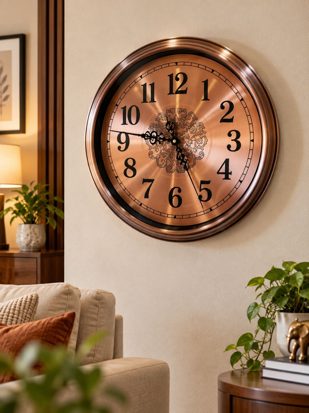 A large round wall clock with a metallic bronze finish and black Arabic numerals, mounted on a beige wall in a living room setting with a sofa and plants visible.