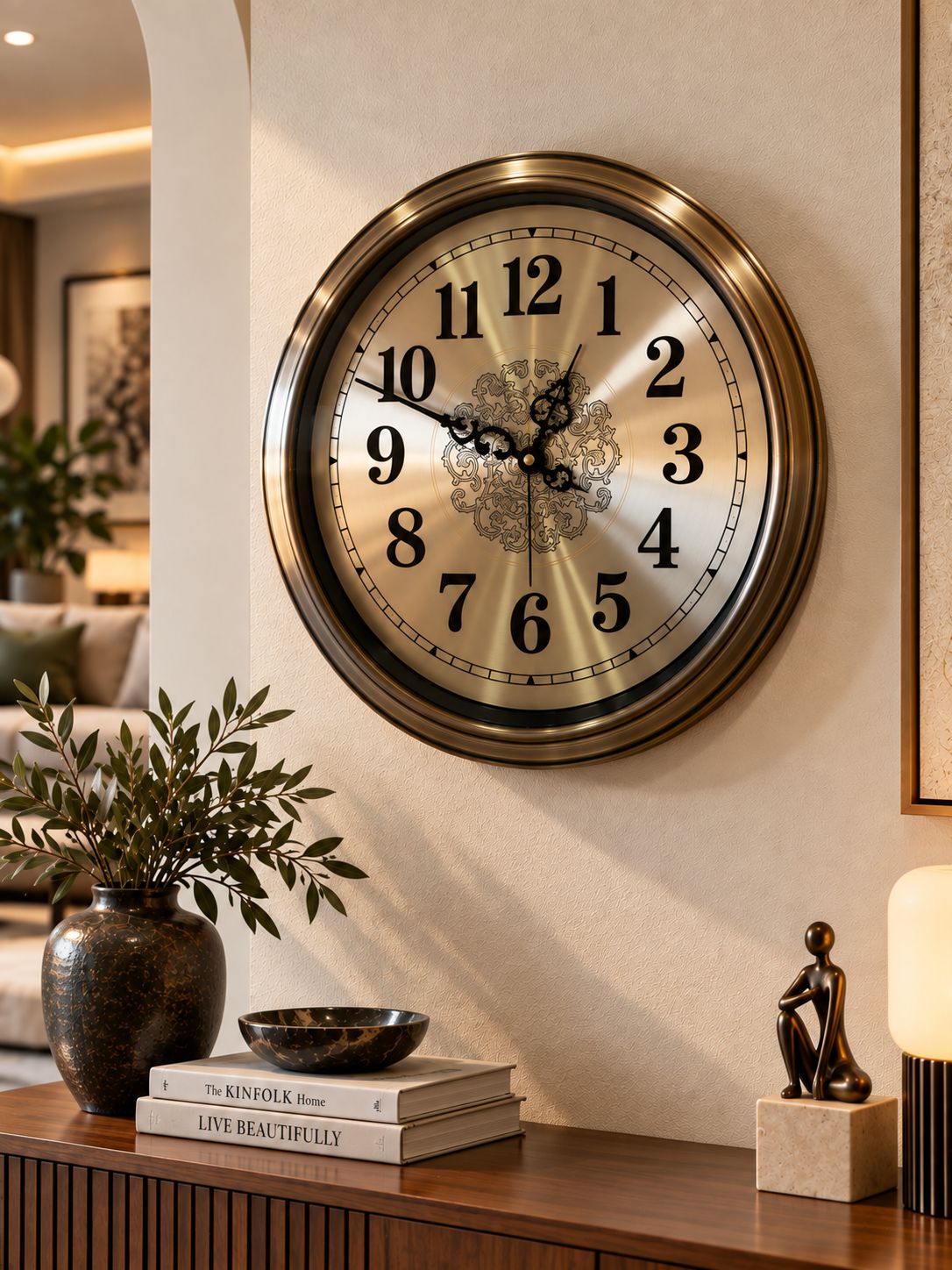 A large round wall clock with a metallic bronze frame and a cream-colored face featuring black Arabic numerals and ornate black hands, mounted on a beige wall in a styled interior setting with books, a plant, and decorative objects below.