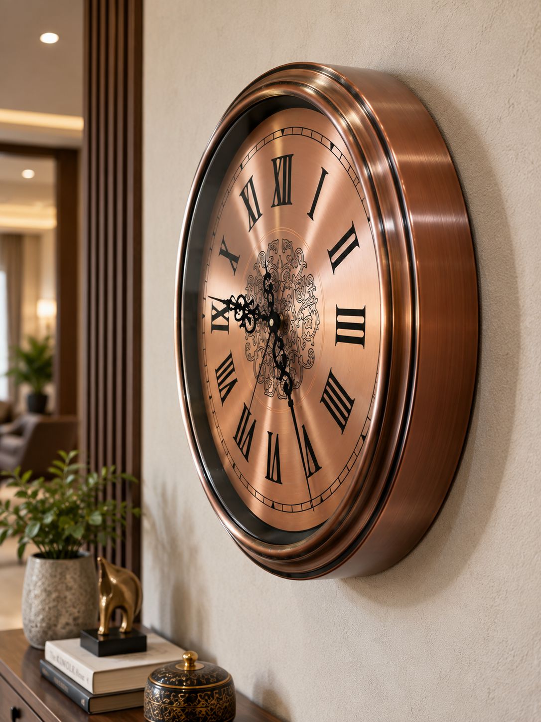 A large round wall clock with a metallic bronze frame and a warm copper-toned face, featuring black Roman numerals and ornate black hands, mounted on a beige wall in a styled interior setting.