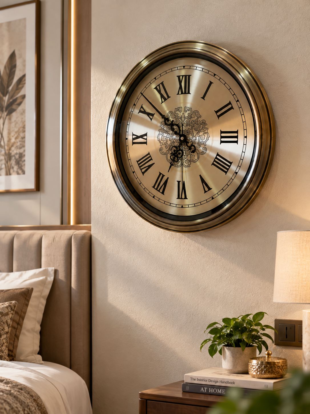 A large round wall clock with a metallic gold frame and Roman numerals, mounted on a beige wall in a styled bedroom setting next to a bed and bedside table.