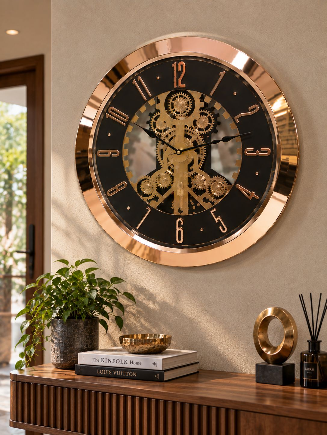 A large round wall clock with a polished rose gold frame and a black face featuring a gold mechanical gear design in the center, mounted on a beige wall above a wooden console table with a plant and decorative objects.