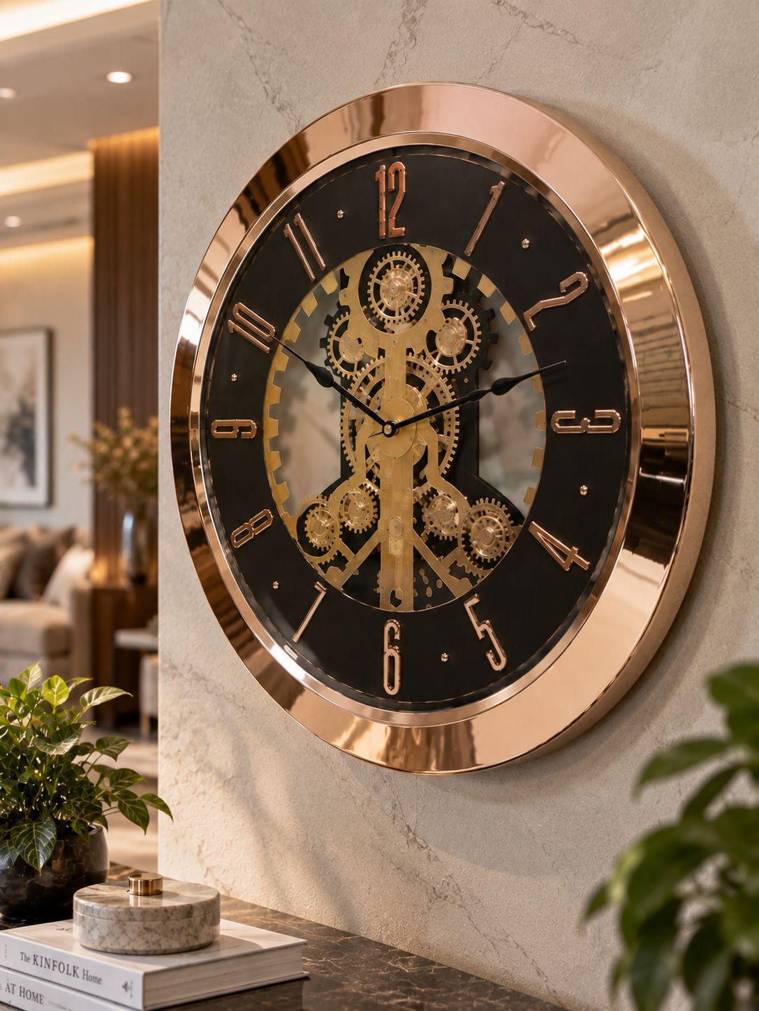 A large round wall clock with a polished rose gold frame and a black face featuring a gold mechanical gear design, mounted on a light marble wall in a modern living room setting.