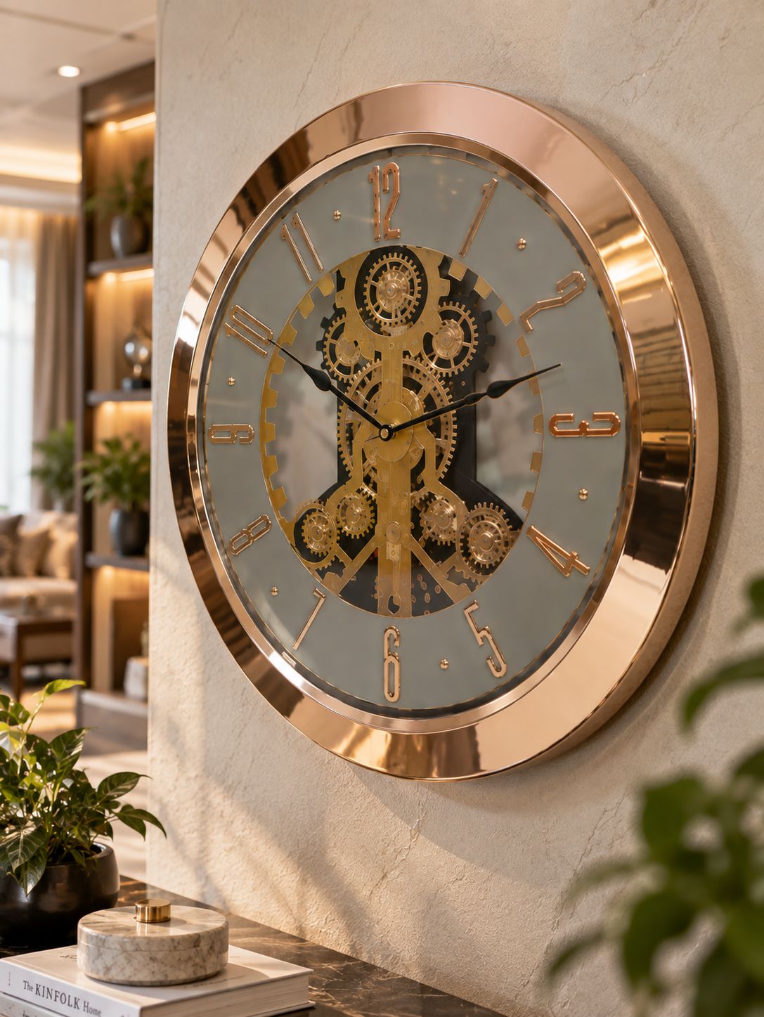 A large round wall clock with a rose gold metallic frame and a detailed gold mechanical gear design on the face, mounted on a textured beige wall in a modern living room setting.